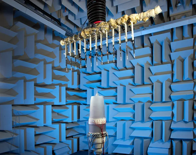 Microphones and suspended testing apparatus inside a chamber lined with blue acoustic wedges.