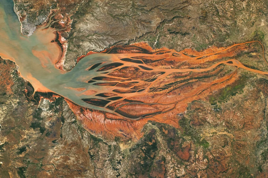 Rust-red sediment channels branch through the green and brown landscape of Madagascar’s Betsiboka River delta.