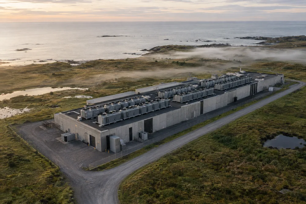 Drone view of a long, low data facility with rows of rooftop cooling units on a coastal plain, fog drifting over the grassland and the sea beyond.