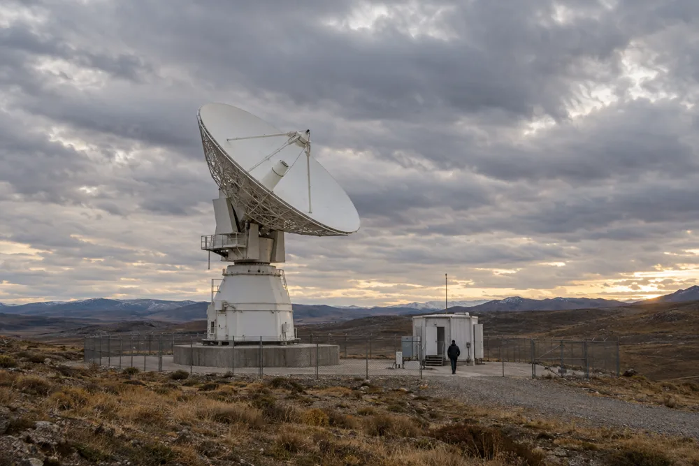 A large white satellite ground-station dish inside a perimeter fence under a heavy evening sky, a small service hut beside it and a person walking across the gravel.
